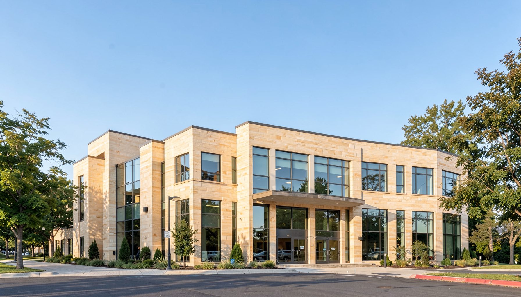A wide, softly lit view of a modern medical office building exterior with warm beige stone and large windows, set against a clear blue sky with gentle tree-lined surroundings